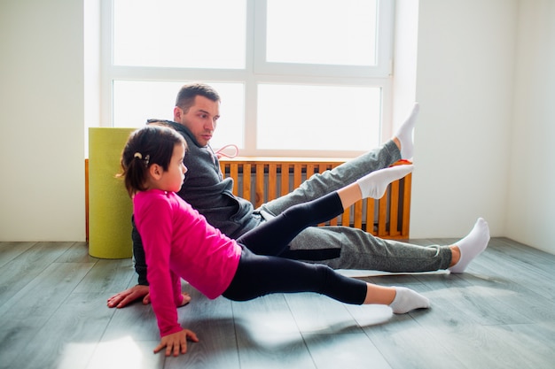 Young father and his cute little daughter are doing reverce plank with leg raise on the floor at home. Family fitness workout. Cute kid and daddy is training on a mat indoor near the window in room.