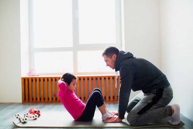 Young father and his cute little daughter have ABS workout at home. Cute kid and daddy is training on a mat indoor. Little dark-haired female model in sportswear has exercises near the window in room