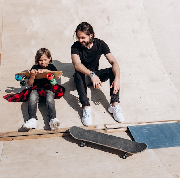 스타일리시한 캐주얼 옷을 입은 젊은 아버지와 그의 아들이 화창한 날씨에 스케이트 파크의 스케이트보드 옆 미끄럼틀에 함께 앉아 있습니다.