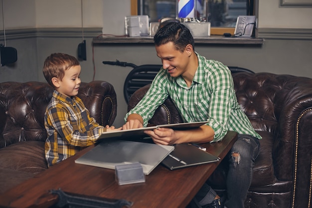 Young father and his stylish little son in the barbershop in the waiting room