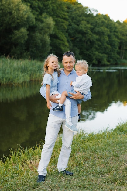 Молодой отец держит своего сына и дочь на руках летом в парке возле озера