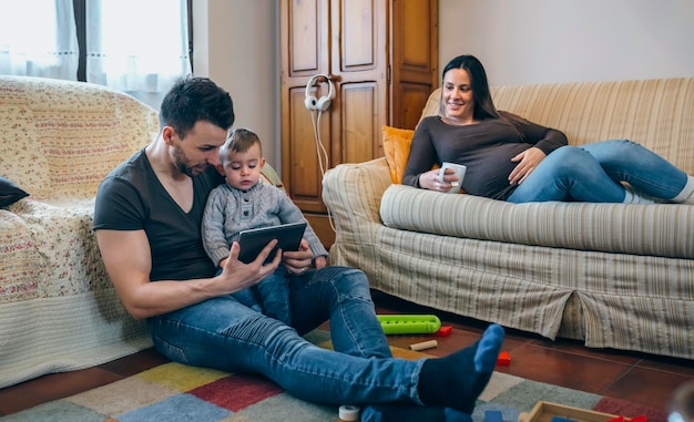 Young father looking at the tablet with his little son while the pregnant mother takes a tea lying on the couch