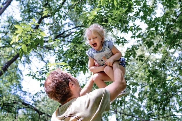 Young father raises his little cheerful daughter high up with his hands against the sky and green trees