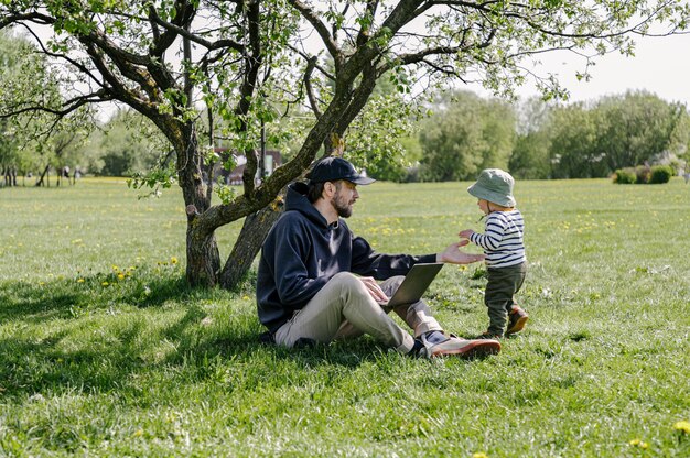 young father sits with a laptop in the park working and playing with a child