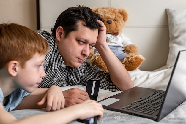 Young father and son shopping online together family time Child redhaired boy and dad at the laptop choose purchases The man is holding a debit card