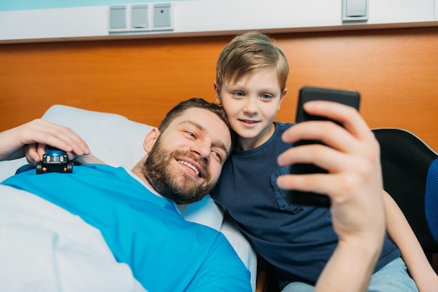 Young father and son taking selfie on smartphone at ward hospital patient care