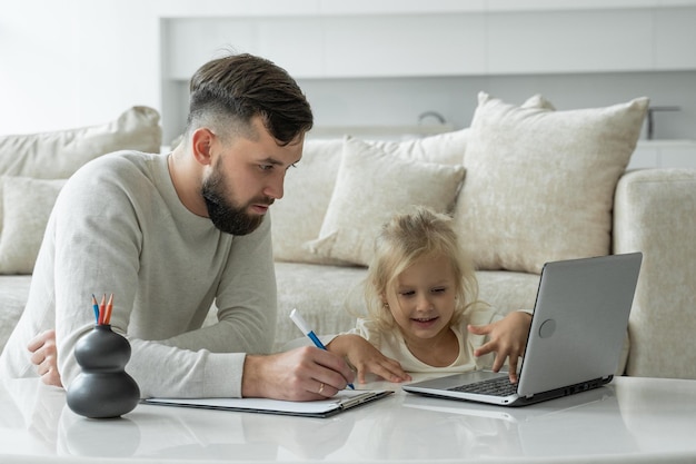 Young father teaches a young daughter using a laptop near the sofa in the living room of the house