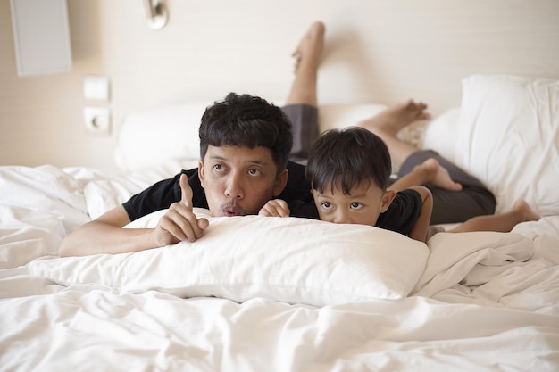 Young father and toddler boy playing on bed cheerfully in hotel room on vacation