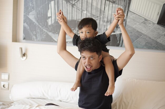 Young father and toddler boy playing on bed cheerfully in hotel room on vacation