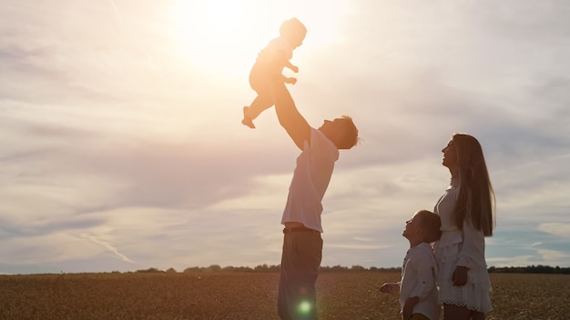 Young father tosses toddler son and mother in white dress with elder boy looks among yellow wheat field, sunlight