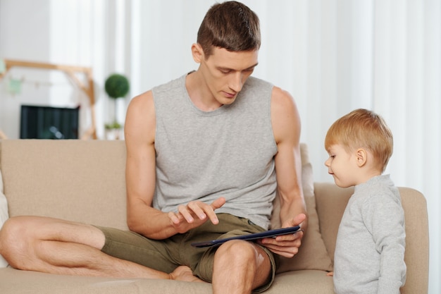 Young father turning on the cartoons on digital tablet for his little son at home