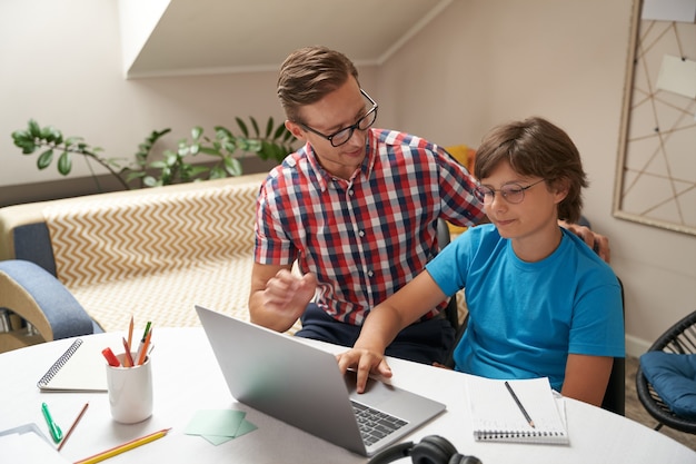 Foto giovane padre che indossa occhiali aiutando suo figlio a fare i compiti ragazzo usando il laptop