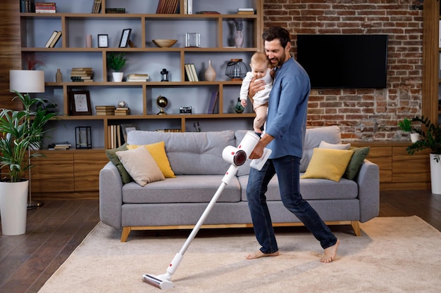 A young father with a child does housework Father on maternity leave Dad cheerfully sings dances and vacuums cleans with a small child in his arms Paternity Leave