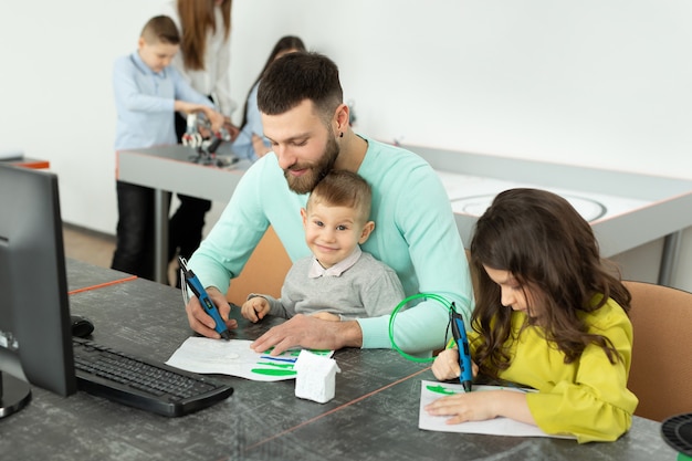 Молодой отец с сыном и дочерью рисуют рисунок 3D ручкой на уроках робототехники
