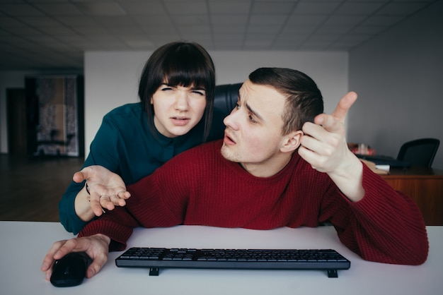 Young fellow man and woman confused looking at the computer monitor. Hipster in a modern office.