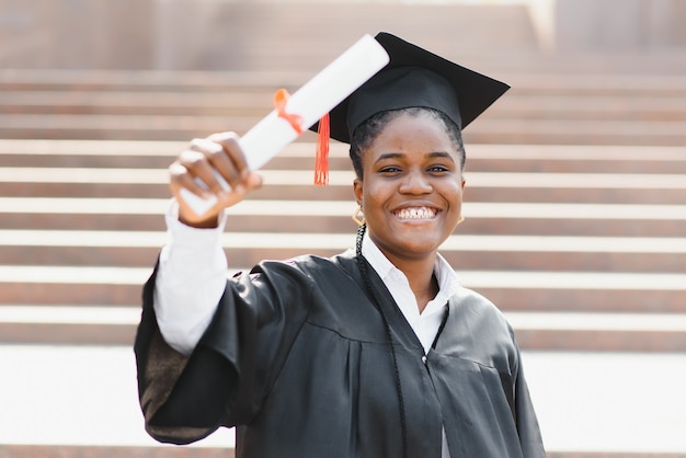 卒業証書を持つ若い女性のアフリカ系アメリカ人の学生は、屋外でポーズをとる。