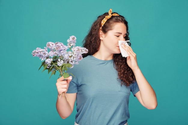 Young female allergic to blooming lilac blowing nose into paper handkerchief while standing over blue wall in isolation