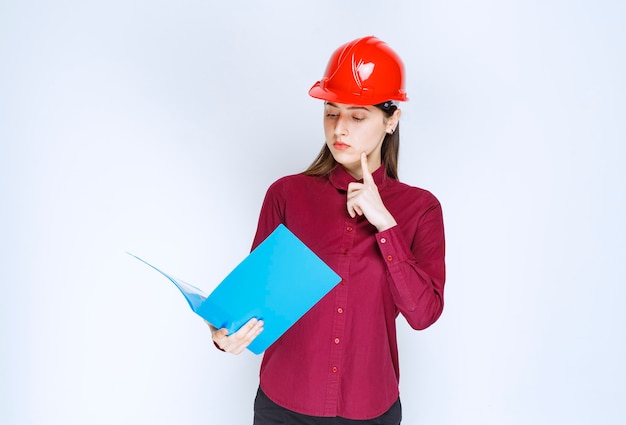 Фото Молодой женский архитектор в красном шлеме, читая важные примечания на белом фоне.