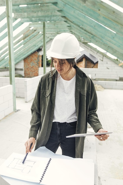 Photo young female architect with tablet checking blueprints against wooden roof framing of modern farmhouse stylish woman engineer in hard hat looking at digital plans at construction site