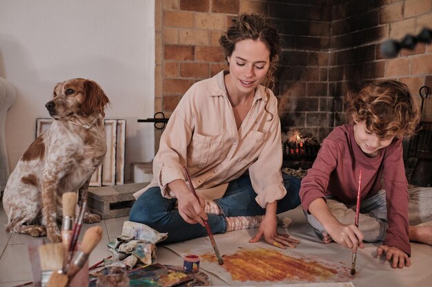 Photo young female artist in casual clothes and boy with brush painting on paper and looking down while spending time together at home near spaniel dog