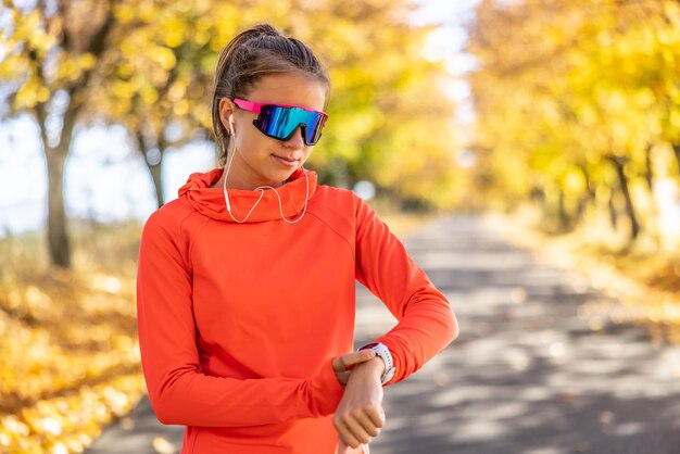 A young female athlete checks her running performance on a smart watch listens to music through headphones and wears sports sunglasses