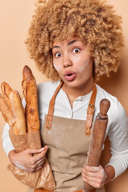 Young female baker holds paper bag of baguettes and rolling pin\
wears brown apron bakes fresh homemade bread eats delicious natural\
products has amazed expression isolated over beige background