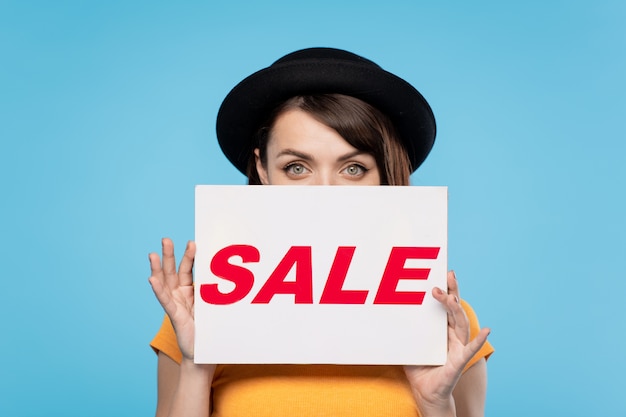 Молодая женщина в черной шляпе выглядывает из бумажного плаката с объявлением сезонной распродажи изолированно