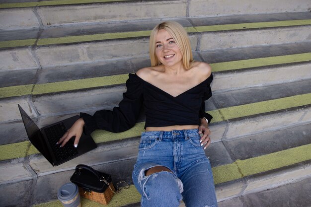 A young female blogger is sitting on the steps and working on a laptop