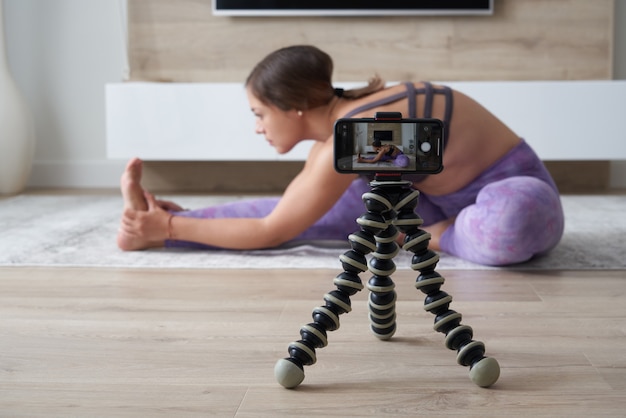 Young female blogger recording sports video at home doing yoga in living room