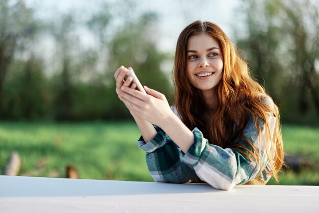 若い女性ブロガーがスマートフォンを手に屋外に座り、屋外でフリーランスをしている健康的なライフスタイルと仕事のコンセプト