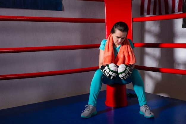 Young female boxer with a towel around her neck sitting in regular boxing ring in a gym