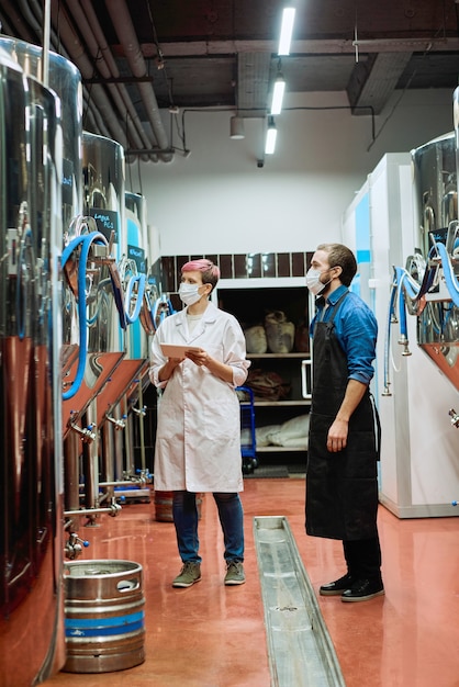 Photo young female brewery expert in whitecoat and protective mask and male brewer standing in aisle in front of new beer production equipment