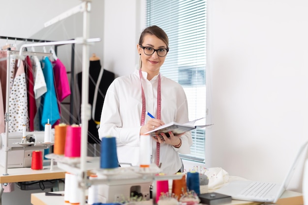 Young female business owner takes notes into her notepad while standing in her tailor workshop