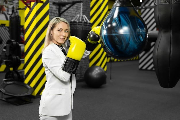 Young female businesswoman in a white suit hits a punching bag in the gym. The concept of office employee anger and relaxation.