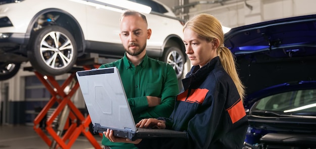 A young female car mechanic holds a diagnostic laptop and shows the manager the identified errors against the background of the car Panoramic photography