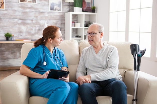 Young female caretaker sitting on couch with senior man in nursing home.