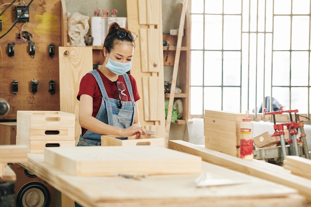 Young female carpenter in protective mask polishing drawer with wooden block when working at workbench