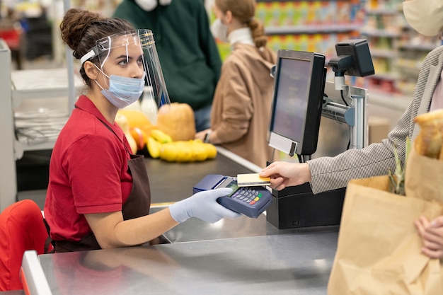 Giovane cassiera in uniforme e maschera protettiva, guanti e schermo che guarda uno dei consumatori che pagano le merci con carta di credito