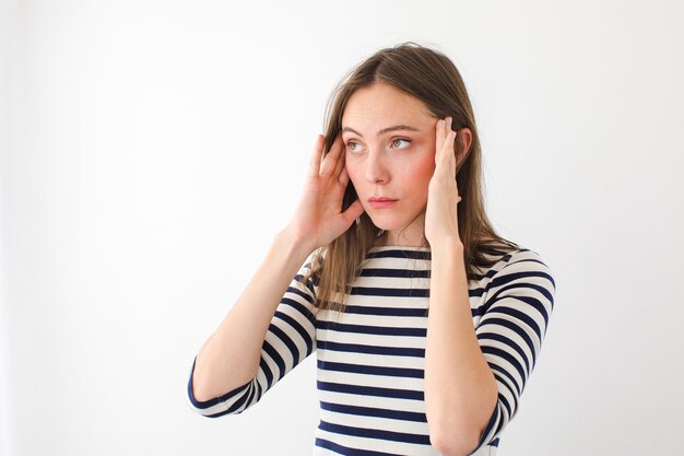 Young female in casual striped wear having headache and rubbing temples while standing against white background