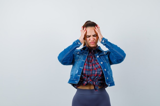 Young female in checkered shirt, jacket, pants with hands on head and looking fatigued , front view.