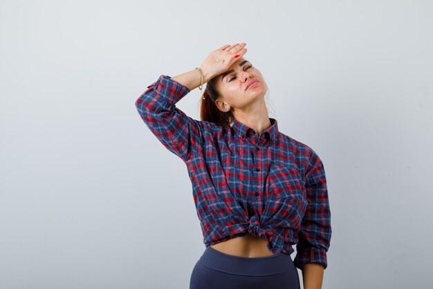 Giovane donna in camicia a scacchi, pantaloni con la mano sulla fronte e sguardo esausto, vista frontale.