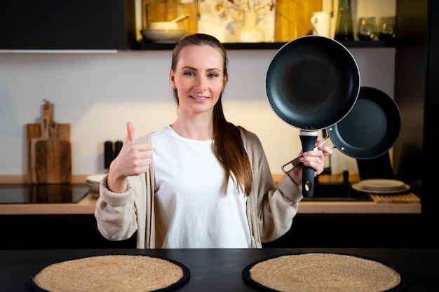 A young female chef holds two frying pans against the background of her kitchen