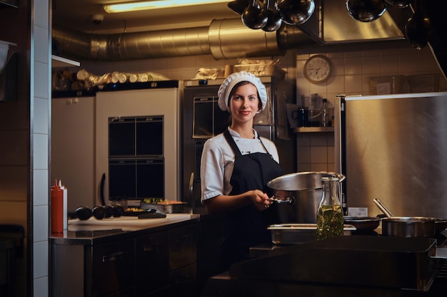Молодая женщина-повар в униформе держит кастрюлю, улыбаясь, глядя в камеру на кухне.