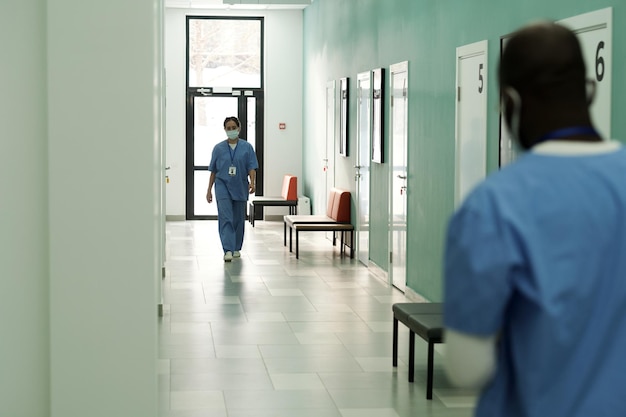 Young female clinician in blue uniform and mask walking along hospital corridor