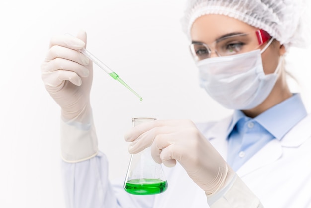 Молодая женщина-врач или химик в защитной спецодежде капает жидкое вещество в клюв с зеленой жидкостью