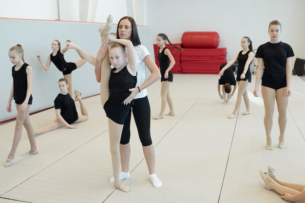 Фото Молодая женщина-тренер помогает гибкой девушке делать шпагат, стоя на одной ноге, в то время как другие девушки выполняют упражнения на разминку