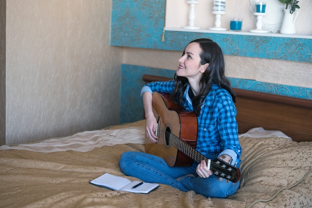 Photo young female composer composes a song on acoustic guitar on bed in bedroom