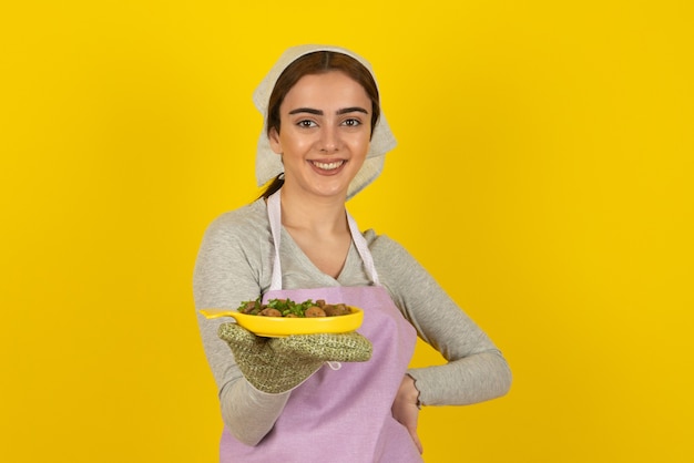 Молодая женщина-повар в фиолетовом фартуке позирует с тарелкой жареных грибов над желтой стеной.