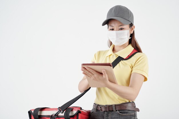 Young female courier in medical mask checking orders via application on tablet computer