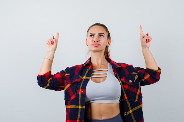 Young female in crop top, checkered shirt, pants pointing up and looking pensive , front view.